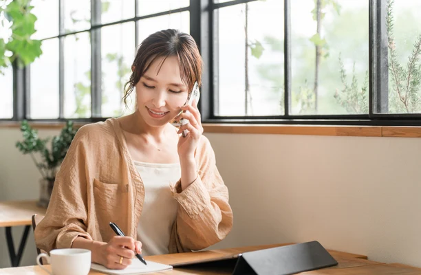 自宅の窓辺で電話をしながらメモを取る社員